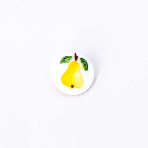 May include: A white ceramic button with a yellow pear and green leaves painted on it.