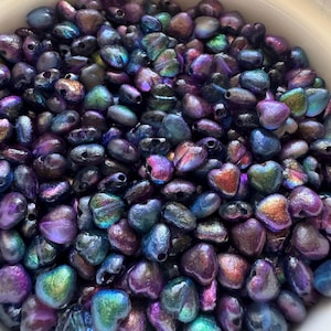 May include: A bowl filled with iridescent heart-shaped beads. The beads are a mix of purple, blue, and green colors with a metallic sheen.