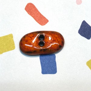 以下が含まれることがあります： 2つの穴がある楕円形のボタン。ボタンは、光沢のある琥珀色の表面に、テクスチャ加工されたまだら模様があります。ボタンの穴は黒色です。ボタンは、カラフルな幾何学模様のある白い背景にあります。