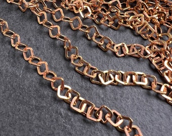May include: Close-up of several strands of copper-colored chain. The chain links are square-shaped with rounded corners. The chains are arranged in a slightly overlapping pattern against a dark gray background, creating a textured appearance.