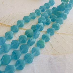 May include: A strand of light blue glass beads with a shell-like design. The beads are arranged in a row and are strung on a clear thread.