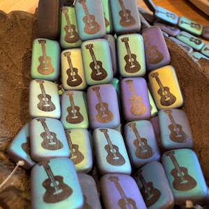 May include: A collection of iridescent glass guitar-shaped beads. The beads are rectangular and have a black silhouette of a guitar on them. The beads are in various shades of blue, green, and purple.