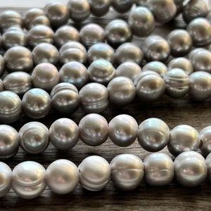 May include: A strand of grey pearls, with a subtle iridescent sheen, arranged in a loose pile. The pearls are round and smooth, with a slightly textured surface.