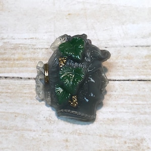 Puede incluir: Un broche de vidrio gris y verde con el perfil de una mujer con hojas y detalles dorados.