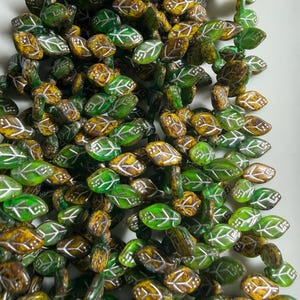 May include: Close-up of leaf-shaped beads in shades of green, brown, and yellow. Each bead features a white leaf vein design. The beads are clustered together, suggesting they are part of a necklace or other jewelry.