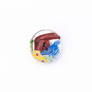 Op de afbeelding: Een doorzichtige glazen knoop met een geschilderd ontwerp van een blauwe vogel voor een bruin vogelhuisje met gele bloemen.