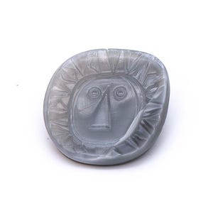 Puede incluir: Un objeto decorativo gris de forma ovalada con un diseño de rostro solar tallado. El sol tiene una cara con ojos, nariz y boca. Los rayos están tallados alrededor de la cara. El objeto está sobre un fondo blanco.