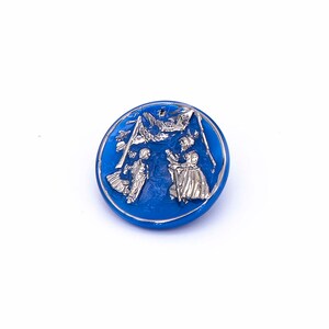 May include: A blue glass button with a silver design of two people skiing in a snowy mountain scene.