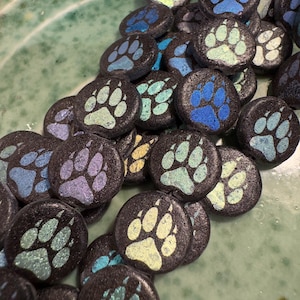 Op de afbeelding: Een verzameling zwarte ronde keramische tegels met een poot afdruk ontwerp in verschillende tinten blauw, groen en paars. De poot afdrukken hebben een glinsterende afwerking.
