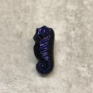 Könnte beinhalten: Ein schwarzer und violetter, irisierender Glas-Seepferdchen-Anhänger. Das Seepferdchen ist mit einem strukturierten Körper und einem eingerollten Schwanz verziert.