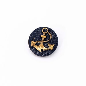 Op de afbeelding: Een ronde, zwarte knoop met een gouden ankerontwerp. Het anker heeft een touwdetail.