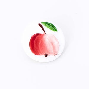 May include: A white ceramic plate with a hand-painted red apple with a green leaf.