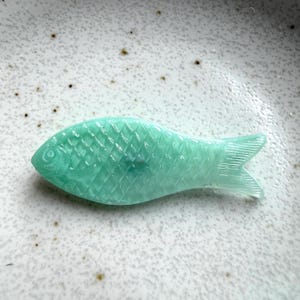 May include: A translucent mint green glass fish figurine featuring a detailed diamond-shaped scale pattern across its body, a defined eye, and a textured tail fin. The object has a smooth, glossy finish and a flat, elongated shape.