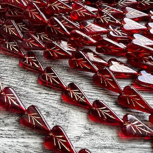 Puede incluir: Una hebra de cuentas de vidrio rojo en forma de corazón con acentos dorados. Las cuentas están dispuestas en filas y tienen una punta puntiaguda.
