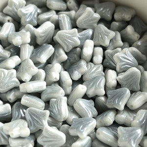 Puede incluir: Primer plano de un montón de cuentas de vidrio grises y blancas. Las cuentas tienen forma de flor con una punta puntiaguda.