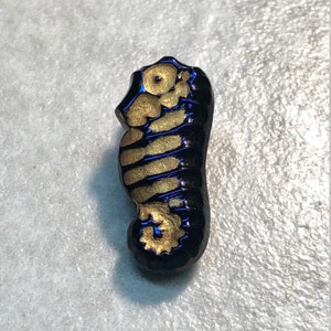 Könnte beinhalten: Ein schwarzer und goldener Glas-Seepferdchen-Anhänger. Das Seepferdchen ist mit goldener Farbe verziert und hat einen schwarzen Hintergrund.