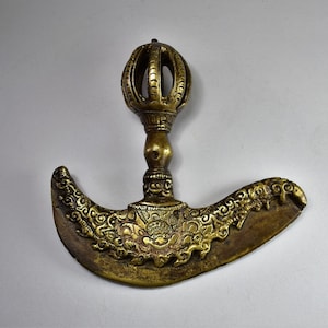 May include: A gold-colored metal ritual weapon with a crescent-shaped blade and a decorative ball-shaped handle. The blade is intricately decorated with floral and geometric patterns.