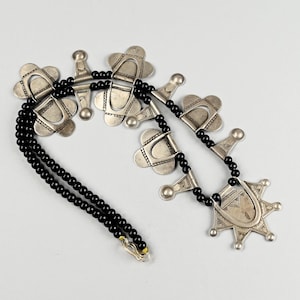 May include: A silver-toned necklace with black beaded strands. The necklace features decorative silver pendants with geometric designs and a starburst-shaped pendant at the bottom. The necklace is displayed on a white background.