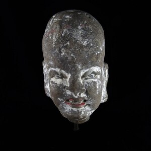 Peut inclure: Une sculpture en pierre grise patinée représentant un visage souriant avec des lèvres peintes en rouge. La sculpture est dépourvue de la partie inférieure du corps et est montée sur un socle noir.