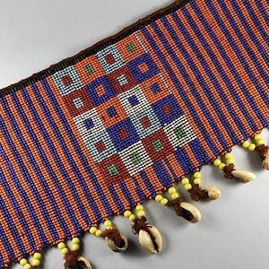 Può includere: Una cintura di perline con un motivo geometrico in tonalità di blu, arancio, rosso e verde. La cintura ha una frangia di perline gialle e conchiglie bianche.