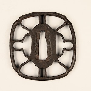 Op de afbeelding: Een vierkante metalen tsuba, een decoratieve bescherming voor een Japans zwaard, met een opengewerkt ontwerp met een centrale ovale opening en vier kleinere openingen.