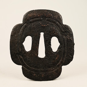 Peut inclure: Une tsuba en métal brun foncé, une garde de sabre japonaise. La garde a une forme symétrique à quatre lobes avec trois ouvertures : deux en forme d'œil et une verticale. La surface a un aspect texturé et vieilli.