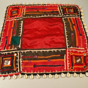 Puede incluir: Una tela cuadrada roja con un borde decorativo de conchas blancas. La tela está decorada con bordados coloridos, incluyendo pequeños espejos y cuentas. El centro de la tela es un cuadrado rojo sólido.