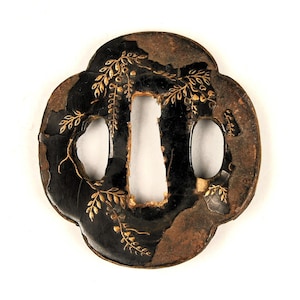 Op de afbeelding: Antieke Japanse tsuba, een zwaardbeschermer, met een donkere achtergrond en een bloemmotief van bladgoud. De tsuba heeft een klaverblad vorm met drie openingen en een centrale sleuf. De randen vertonen tekenen van ouderdom en slijtage.