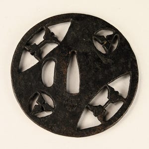 Peut inclure: Une tsuba japonaise circulaire en métal sombre, avec des motifs découpés complexes. La tsuba présente une ouverture centrale et plusieurs formes décoratives, notamment des motifs floraux ou de feuilles stylisés. Le métal a un aspect vieilli.