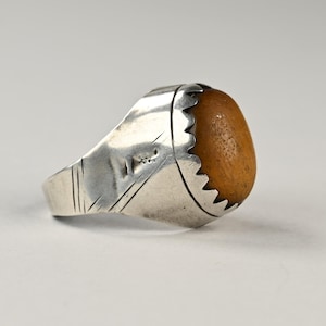 Op de afbeelding: Een zilveren ring met een lichtbruine, ovaalvormige steen. De ring heeft een getextureerde band met geëtste lijnen en een gekartelde bezel setting rond de steen. De ring staat tegen een neutrale achtergrond.