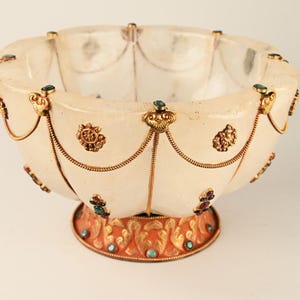 Peut inclure: Un bol décoratif blanc cassé avec des détails dorés et des ornements aux couleurs de bijoux. Le bol présente un bord festonné et est orné de filigranes dorés et de petites pierres précieuses. La base est orange contrastant avec des détails dorés.