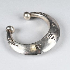 May include: A silver-toned open cuff bracelet with a crescent shape, featuring two rounded ball terminals at the ends. The surface of the metal is decorated with small, stamped patterns of dots arranged in triangular and square groupings.