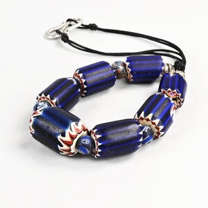 Könnte beinhalten: Ein Armband mit zylindrischen, dunkelblauen Glasperlen mit weißen und roten Chevron-Mustern, durchsetzt mit kleineren, gemusterten Rundperlen. Die Perlen sind auf einer schwarzen Schnur mit einem silberfarbenen Metallhakenverschluss aufgezogen.