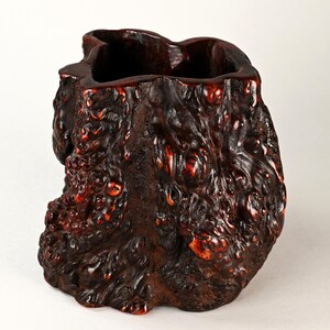 May include: A handcrafted, dark brown wooden vase with a textured, organic surface. The vase has a rough, bark-like appearance with reddish-brown accents. The interior is a dark, smooth cavity.