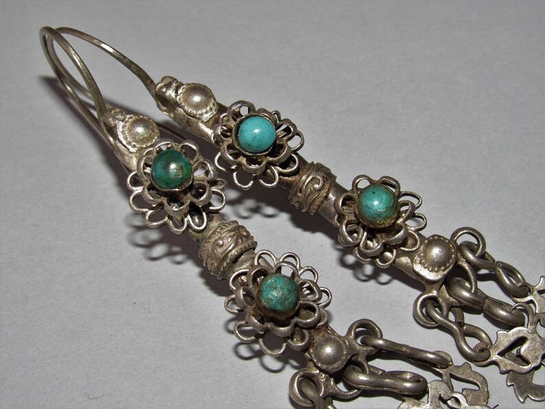 May include: A pair of silver earrings with turquoise stones and intricate floral designs. The earrings have a dangling chain with additional floral accents.