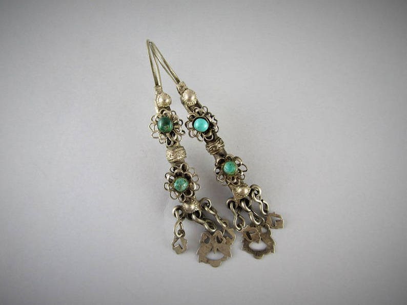 May include: A pair of silver earrings with turquoise stones and intricate filigree designs. The earrings have a dangling charm at the bottom.