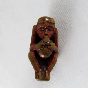 May include: A small wooden figurine of a person sitting with legs crossed, holding a round object in their hands. The figurine is painted red and brown.