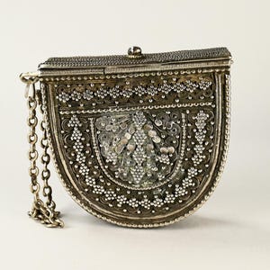 Peut inclure: Un sac à main argenté orné, avec une chaîne. Le sac présente des détails complexes en filigrane et perles, avec un dessus à charnière et un petit fermoir. Le design est symétrique, avec un fond incurvé et un dessus rectangulaire.