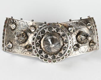 Könnte beinhalten: Aufwendiger silberner Gürtel mit komplizierten floralen und geometrischen Mustern. Der Gürtel hat ein zentrales kreisförmiges Design und mehrere dekorative Elemente. Die Metallarbeiten sind detailliert, mit einer Mischung aus Texturen und kleinen, farbigen Akzenten. Ein Vintage-Accessoire.