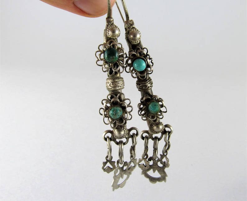 May include: A pair of silver earrings with turquoise stones and intricate filigree designs. The earrings have a dangling charm at the bottom.