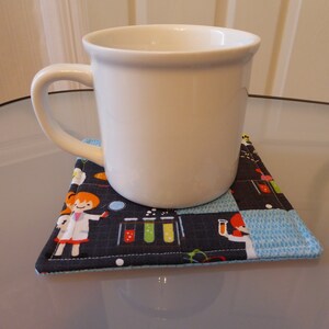 May include: A white ceramic mug sits on a blue and black fabric coaster with a science theme. The coaster features a pattern of test tubes, beakers, and cartoon scientists.