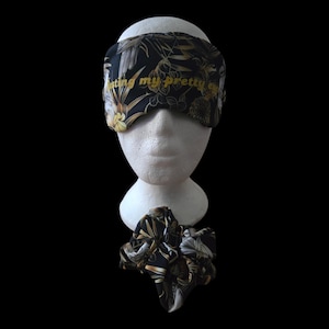 May include: A black sleep mask and matching scrunchie with a gold and white floral pattern. The sleep mask has the text "resting my pretty eyes" in gold lettering. The items are displayed on a white mannequin head against a black background.
