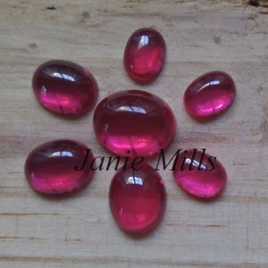 May include: Seven oval-shaped, pink glass cabochons, with a glossy finish, are arranged in a starburst pattern on a wooden surface.