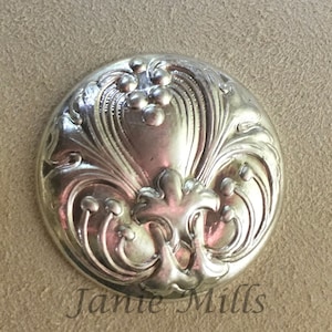 May include: A silver-toned button with an ornate floral design. The button has a raised, three-dimensional design.