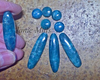 May include: A collection of teal blue gemstone cabochons in various shapes and sizes. The cabochons are arranged in a circular pattern with a few oval shaped cabochons in the center.