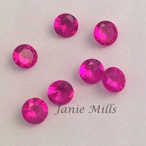 May include: Seven round, pink, faceted gemstones on a white background.  Janie Mills is written in black text below the gemstones.