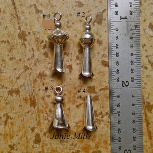 May include: Four silver-coloured metal pendant charms with different designs, including bell-shaped and rounded tops. A ruler is placed next to the charms for scale. The text "Janie Mills" is visible at the bottom.