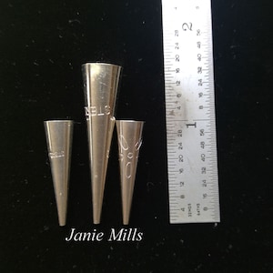 May include: Three silver metal piping tips, the largest is labelled "STER", the middle is labelled "ROELS", and the smallest is labelled "RO" with a flower design. The tips are lined up from largest to smallest, with a ruler in the background.