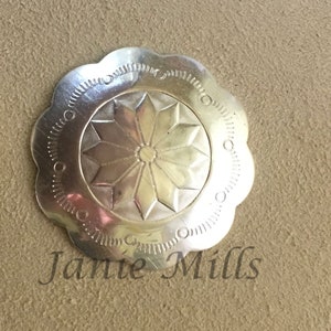 May include: A silver-toned metal flower-shaped button with a starburst design in the center. The button has a scalloped edge and a raised rim around the starburst. The button is on a brown background with the text "Janie Mills" below it.