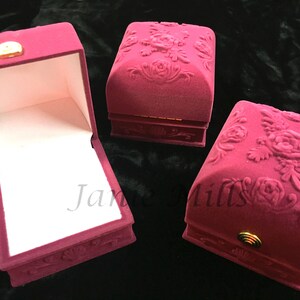 May include: Three small, red velvet ring boxes with a floral design. The boxes are closed, but one is open to show the white velvet interior.